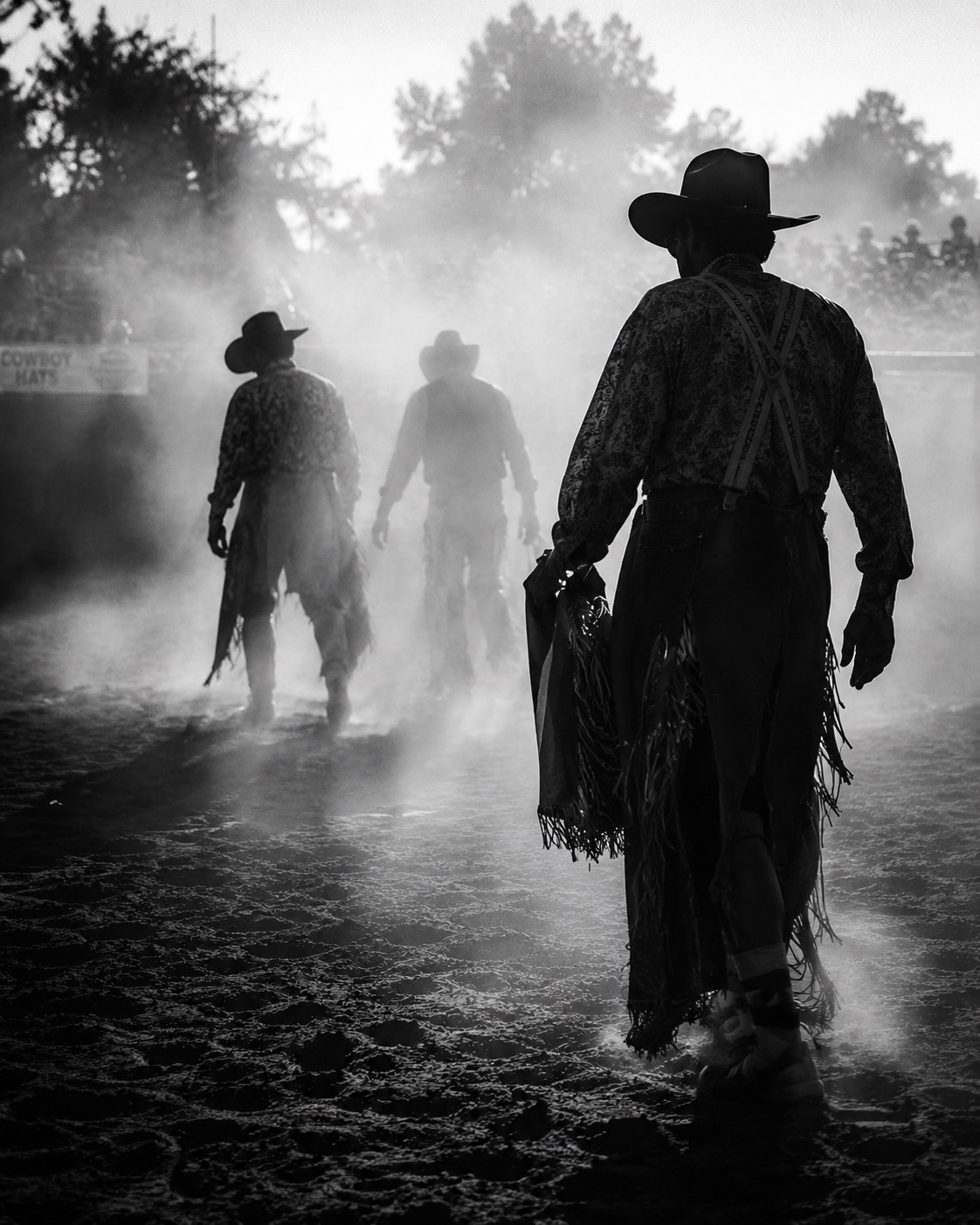 Cowboys walking out backlit through arena dust at dusk — black-and-white western atmosphere by Brian Mullin, Pause Studios.