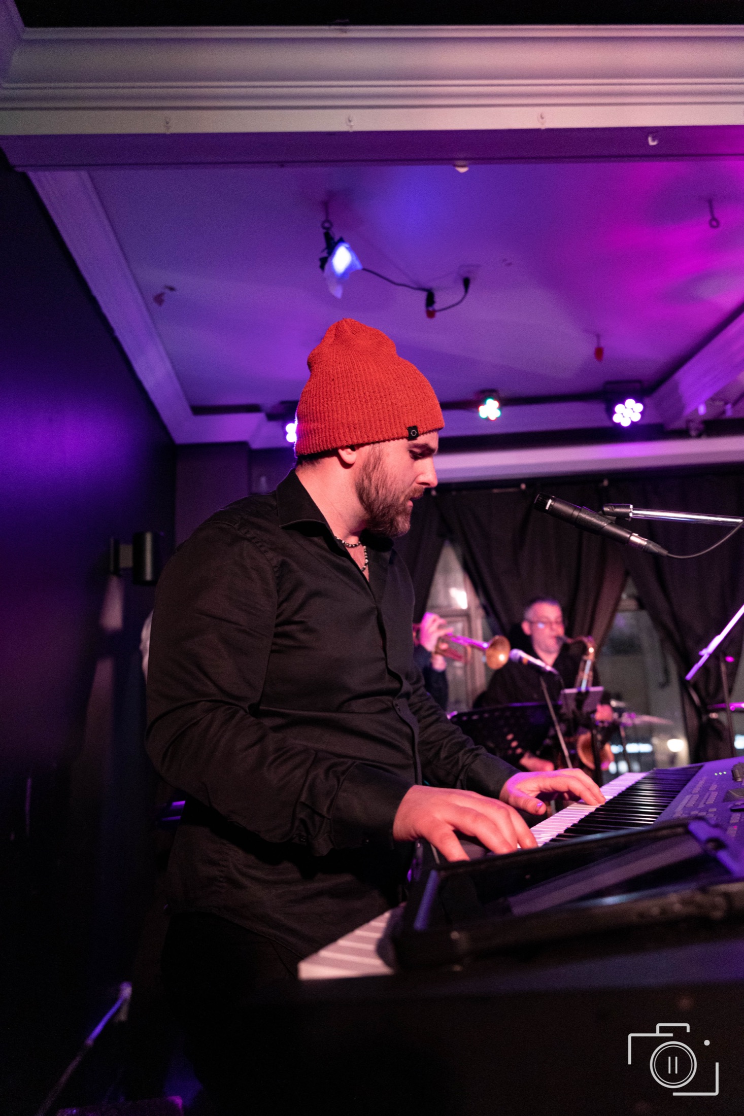 Pianist in red beanie playing keys under purple ambient light, another musician in background — live event photography by Brian Mullin