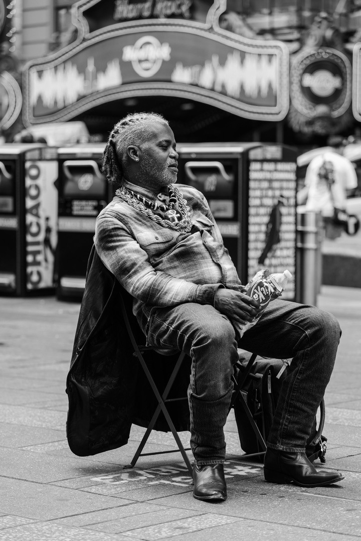 A candid black-and-white street portrait of a man seated in a folding chair outside the Hard Rock in Times Square, New York. Photography by Brian Mullin, Pause Studios.